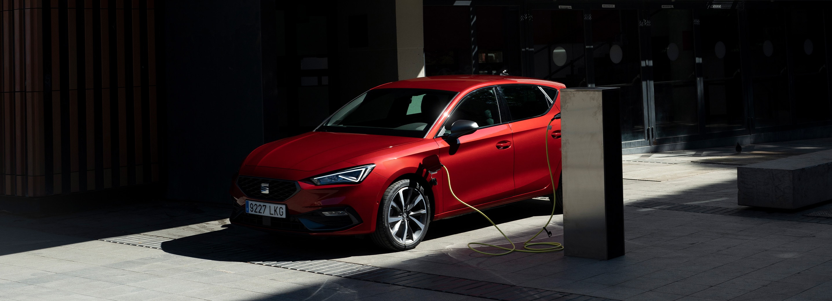 Vue de côté d’une voiture hybride rechargeable SEAT rouge, garée et en train de recharger dans un endroit partiellement ensoleillé et à moitié couvert, mettant en avant l’idée de conduire de manière efficace tout en respectant l’environnement et en réduisant les émissions.