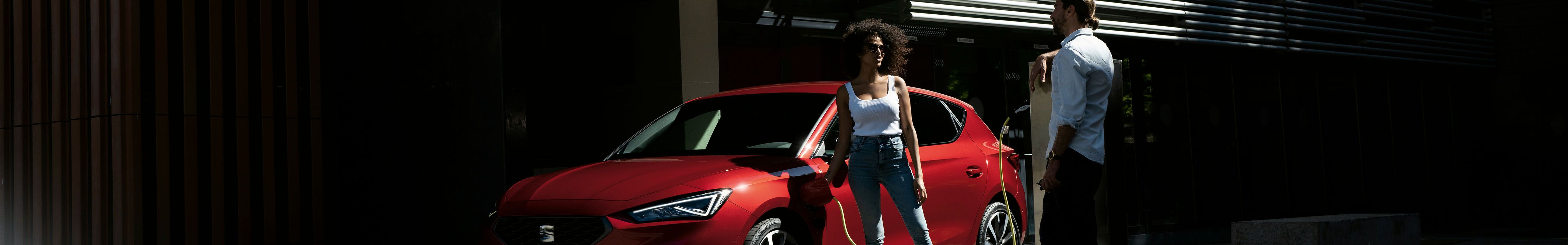 SEAT Leon PHEV rouge, avec un couple discutant de l'opportunité d'acheter une voiture hybride, angle bas et vue de trois quarts avant.